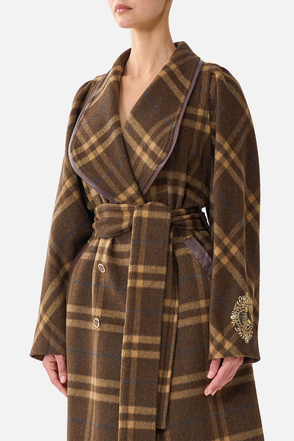 A woman wearing a brown and tan plaid wrap coat with a waist tie, shown from the lower face to the hands against a light gray studio background.